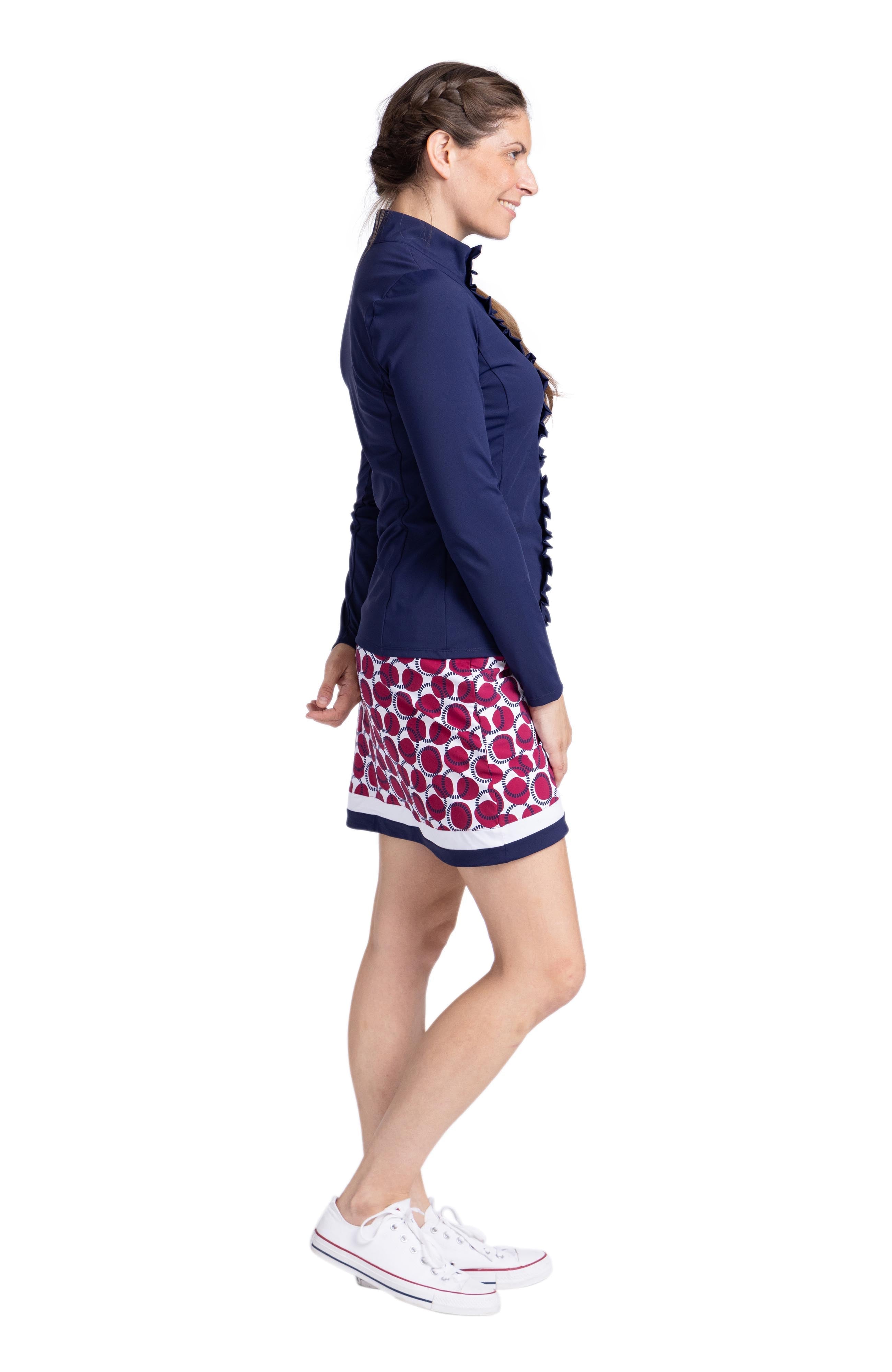 Side view of a woman wearing a navy blue long sleeve top with zipper front closure and ruffle detail down each side of the zipper. The top is worn with a red, white and navy circled pattern skort (Free Drop Skort in Dot and Dash).