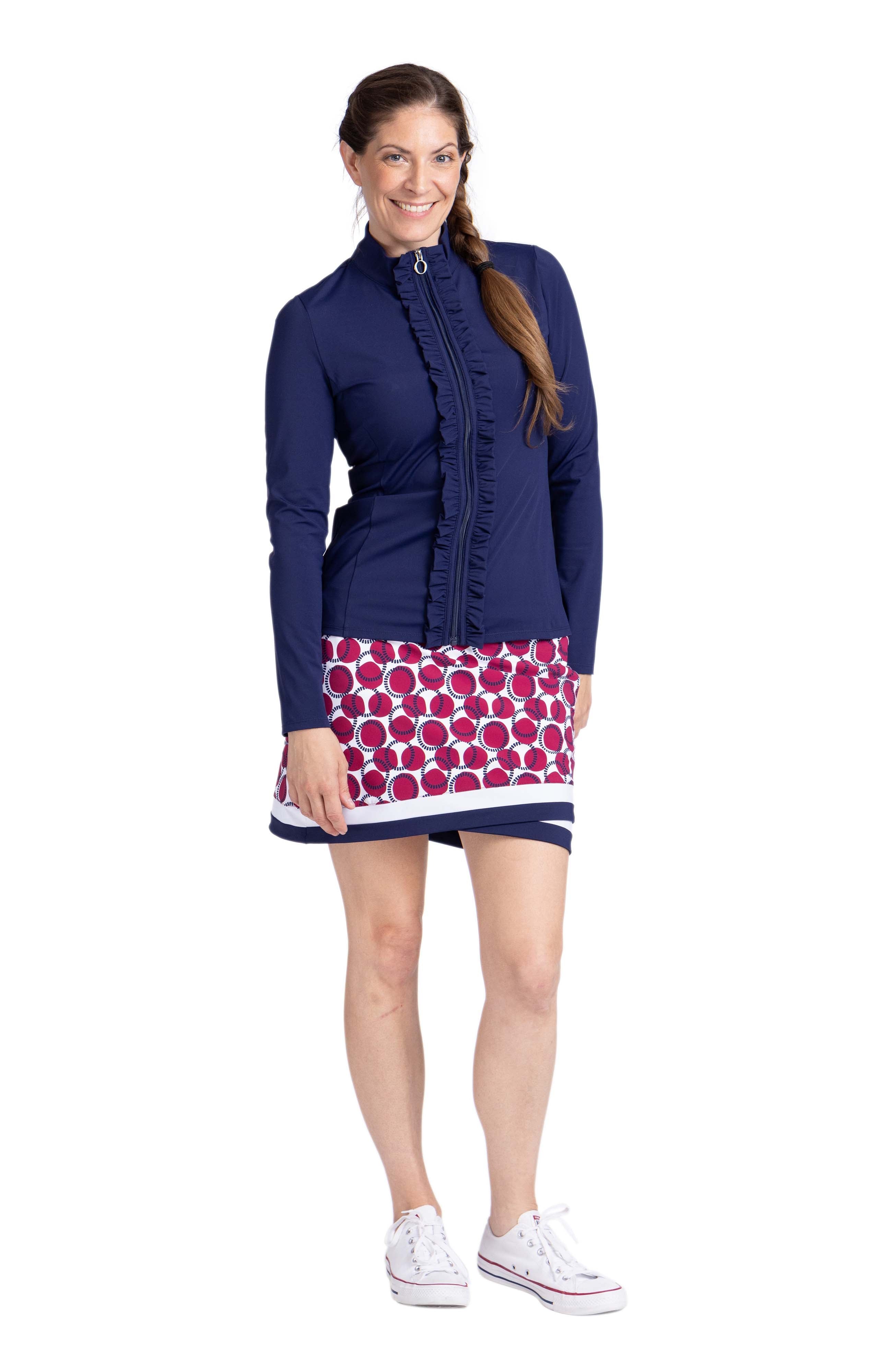 Full view of a woman wearing a navy blue long sleeve top with zipper front closure and ruffle detail down each side of the zipper. The top is worn with a red, white and navy circled pattern skort (Free Drop Skort in Dot and Dash).